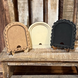 May include: Three small wooden frames in varying colors: light brown, cream, and black. Each frame has an arched top and a scalloped edge. They are displayed on a small wooden table, showcasing their decorative design.