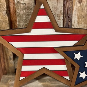 May include: Wooden star-shaped wall decor with a patriotic theme. The large star features red and white stripes, while a smaller star displays a blue background with white stars. Both stars have a brown wooden frame.