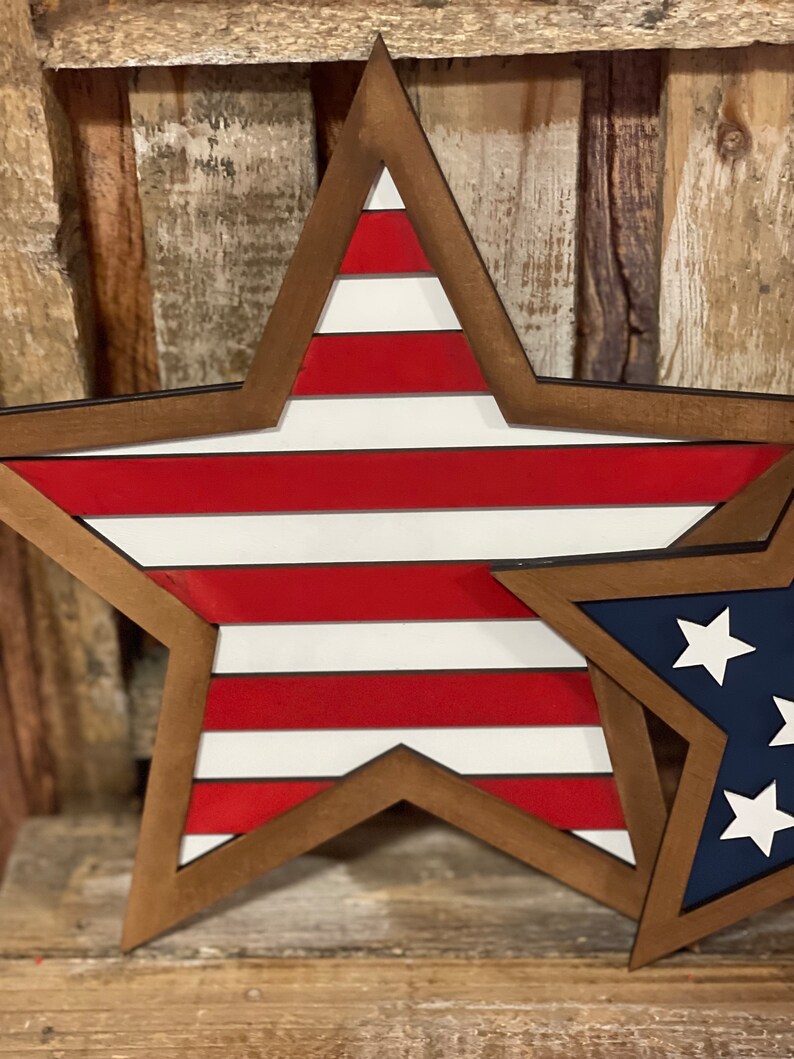 May include: Wooden star-shaped wall decor with a patriotic theme. The large star features red and white stripes, while a smaller star displays a blue background with white stars. Both stars have a brown wooden frame.