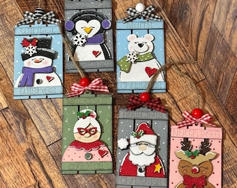 May include: Six wooden Christmas ornaments with different holiday themes. The ornaments are painted in various colors, including red, green, blue, and pink. Each ornament features a different holiday character, including a penguin, a snowman, a bear, Santa Claus, Mrs. Claus, and a reindeer. The ornaments are decorated with glitter, snowflakes, and hearts.