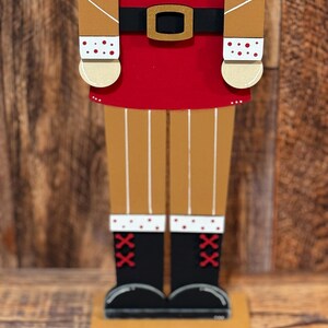 Peut inclure: Une figurine de casse-noisette en bois avec un chapeau ray&eacute; rouge et blanc, un manteau brun, un pantalon rouge et des bottes noires. Le casse-noisette est debout sur une base en bois.