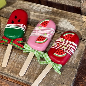 May include: Three wooden popsicle sticks with watermelon designs. The popsicles are decorated with white frosting and green and red sprinkles. The popsicles are tied with green and white gingham ribbon.