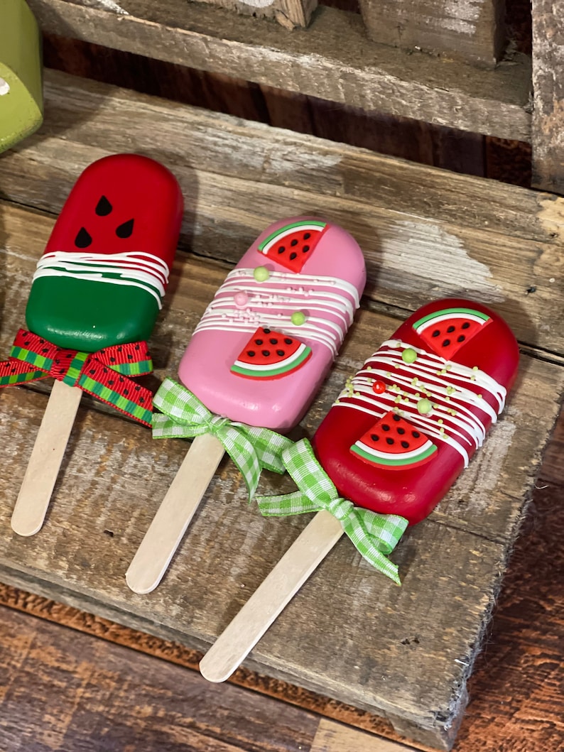 May include: Three wooden popsicle sticks with watermelon designs. The popsicles are decorated with white frosting and green and red sprinkles. The popsicles are tied with green and white gingham ribbon.