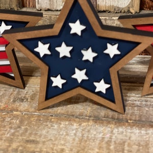 May include: Three wooden stars painted in the colours of the American flag. The stars are blue with white stars and have a brown wooden frame.