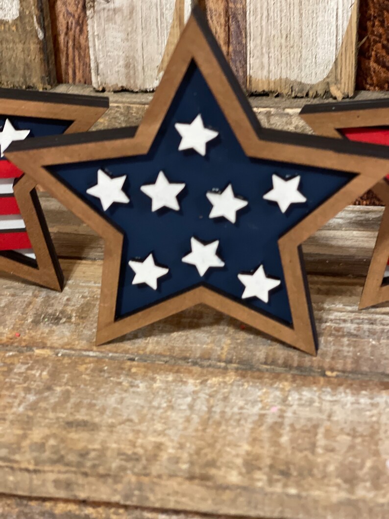 May include: Three wooden stars painted in the colours of the American flag. The stars are blue with white stars and have a brown wooden frame.