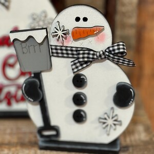 May include: A white wooden snowman figurine with black eyes, a carrot nose, and a black and white checkered ribbon. The snowman is holding a gray bucket with the word "Brrr" written on it. The snowman is decorated with black buttons and white snowflakes.