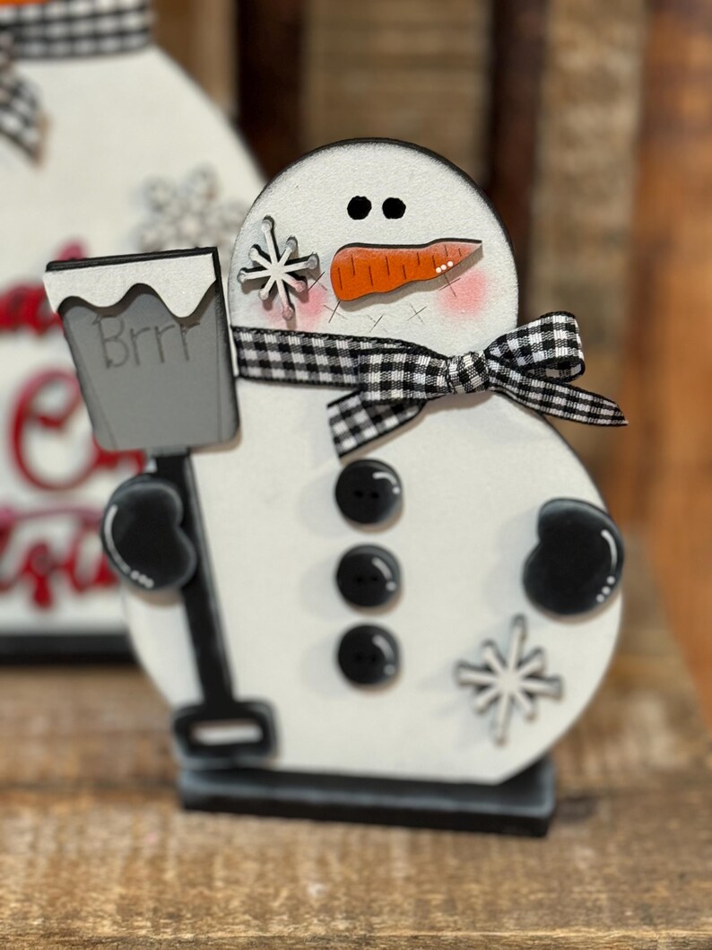 May include: A white wooden snowman figurine with black eyes, a carrot nose, and a black and white checkered ribbon. The snowman is holding a gray bucket with the word "Brrr" written on it. The snowman is decorated with black buttons and white snowflakes.