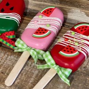 May include: Three watermelon-themed cake pops on wooden sticks. The cake pops are decorated with white chocolate, sprinkles, and watermelon slices. The sticks are tied with green and white gingham ribbon.