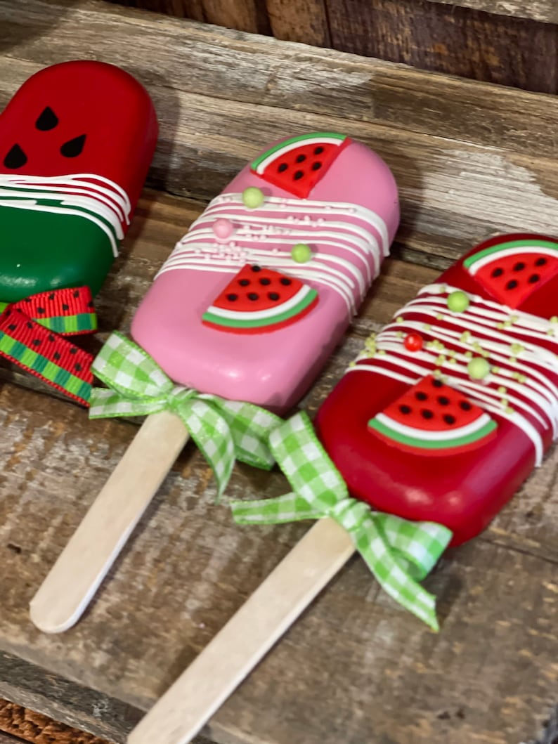 May include: Three watermelon-themed cake pops on wooden sticks. The cake pops are decorated with white chocolate, sprinkles, and watermelon slices. The sticks are tied with green and white gingham ribbon.