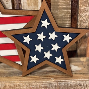 May include: Wooden star-shaped decoration with a dark brown frame, navy blue interior, and white stars. Another decoration with red and white stripes is partially visible. The decorations are placed on a wooden surface.