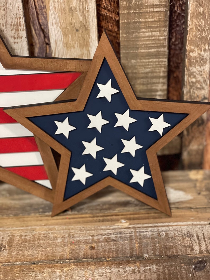 May include: Wooden star-shaped decoration with a dark brown frame, navy blue interior, and white stars. Another decoration with red and white stripes is partially visible. The decorations are placed on a wooden surface.
