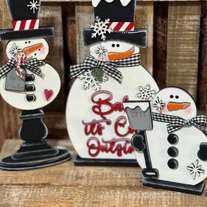 May include: Three wooden snowman figurines with black top hats, black buttons, and orange carrot noses. The middle snowman has a sign that reads "Baby it's Cold Outside".
