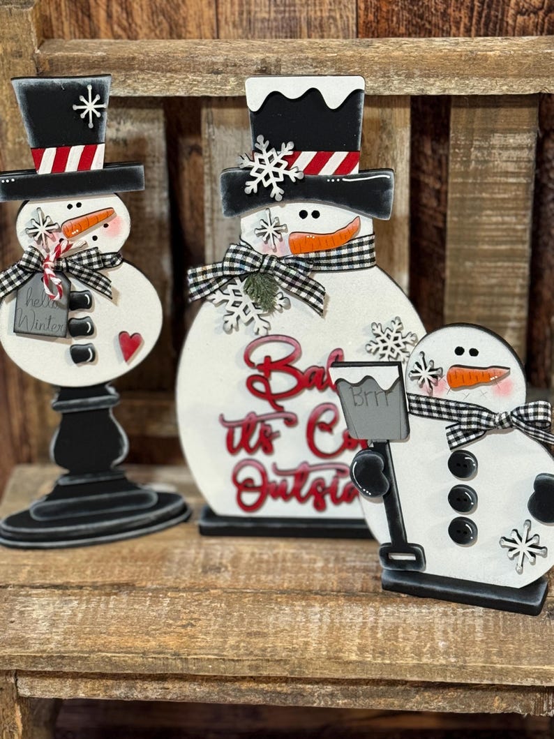 May include: Three wooden snowman figurines with black top hats, black buttons, and orange carrot noses. The middle snowman has a sign that reads "Baby it's Cold Outside".