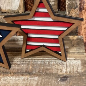 May include: A wooden star with a red, white and blue striped design. The star is painted with a black outline and has a rustic wood grain texture.