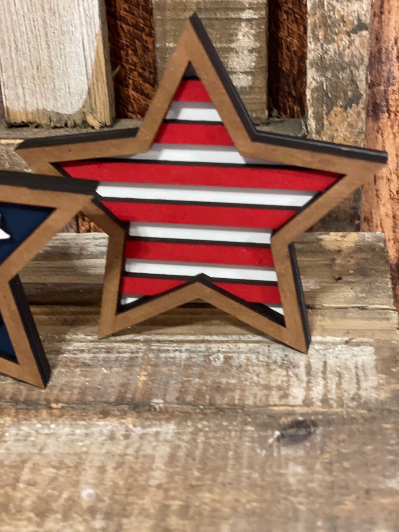 May include: A wooden star with a red, white and blue striped design. The star is painted with a black outline and has a rustic wood grain texture.