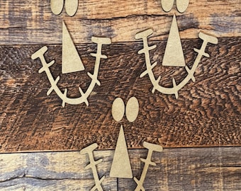 May include: Wooden craft pieces arranged to form three smiling faces. Each face includes oval eyes, a triangular nose, and a curved mouth with spikes. The pieces are light brown and set against a wood grain background.