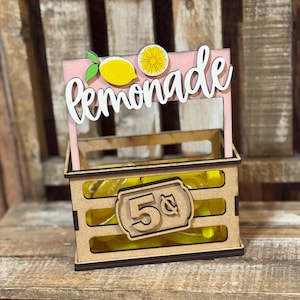 May include: A wooden crate-style display with a pink sign featuring the word lemonade in white script, decorated with yellow lemon graphics. The front of the crate has a rectangular plaque with the text 5 cents, filled with yellow items.