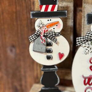 May include: A wooden snowman figurine with a black top hat, red and white striped hat band, black buttons, a carrot nose, and a red heart. The snowman is holding a candy cane and a tag that says "hello winter".