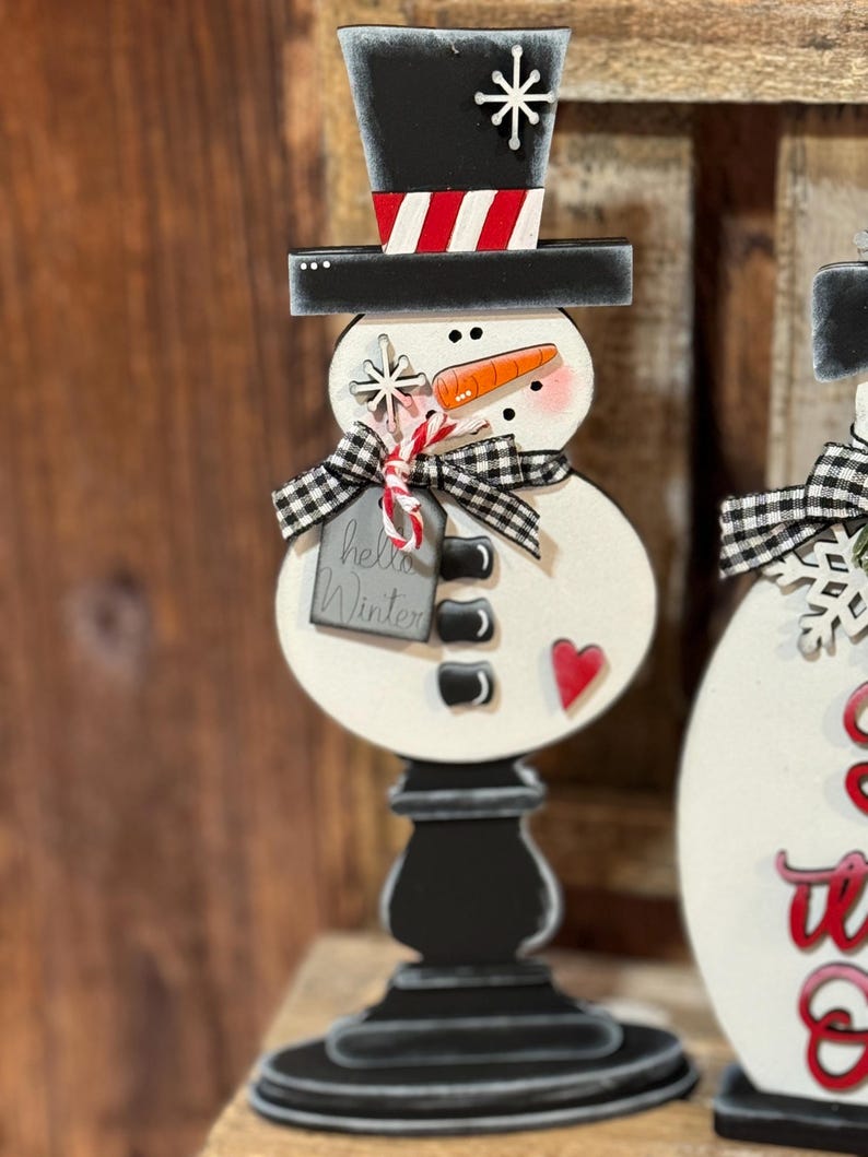 May include: A wooden snowman figurine with a black top hat, red and white striped hat band, black buttons, a carrot nose, and a red heart. The snowman is holding a candy cane and a tag that says "hello winter".