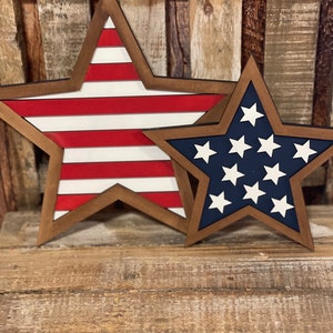 May include: Two wooden star-shaped decorations. One star features red and white stripes, while the other has a blue background with white stars. Both have a brown border and are placed on a wooden surface.