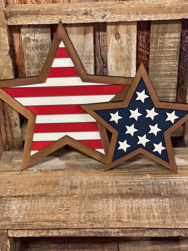 May include: Two wooden star-shaped decorations. One star features red and white stripes, while the other has a blue background with white stars. Both have a brown border and are placed on a wooden surface.