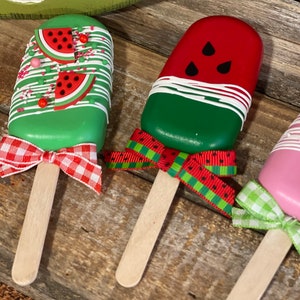 May include: Three watermelon popsicle crafts with wooden sticks and red and green gingham ribbon bows. The popsicles are painted to look like watermelon with red, green, and black paint.