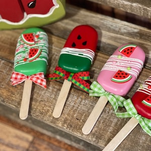 May include: Four wooden popsicle sticks decorated with watermelon designs. Each popsicle has a different color base, including green, red, pink, and a combination of red and gold. The popsicles are tied with a ribbon bow in various colors, including red, green, and white.