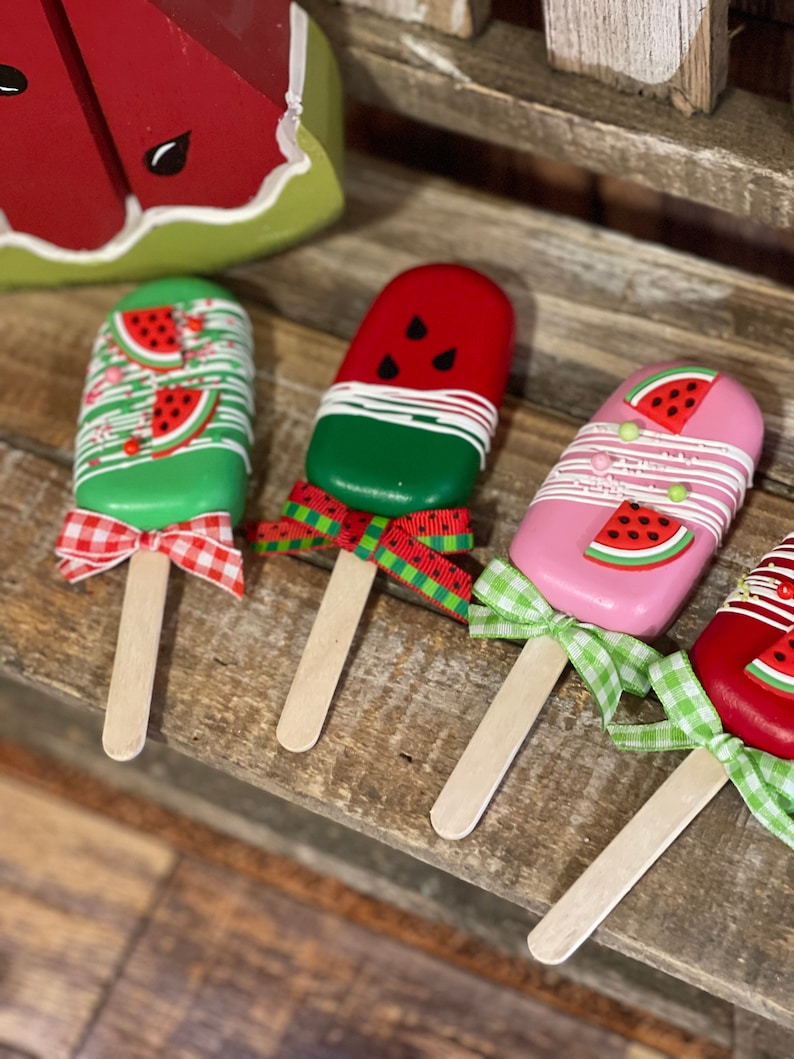 May include: Four wooden popsicle sticks decorated with watermelon designs. Each popsicle has a different color base, including green, red, pink, and a combination of red and gold. The popsicles are tied with a ribbon bow in various colors, including red, green, and white.