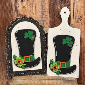 May include: Two St. Patrick's Day themed wooden decorations. One is a scalloped-edge arch, the other a cutting board shape. Both feature a black leprechaun hat with a green belt, orange buckle, and shamrock accents. The background is white with red polka dots.