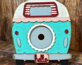 May include: A decorative wooden camper in turquoise, white, and red. The camper features a scalloped edge, a round opening, and the word "hello" on the bumper. The camper has black wheels and is set against a wooden background.