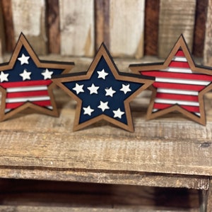 May include: Three wooden stars with a red, white and blue American flag design. The stars are painted in a rustic style and have a brown wood finish.