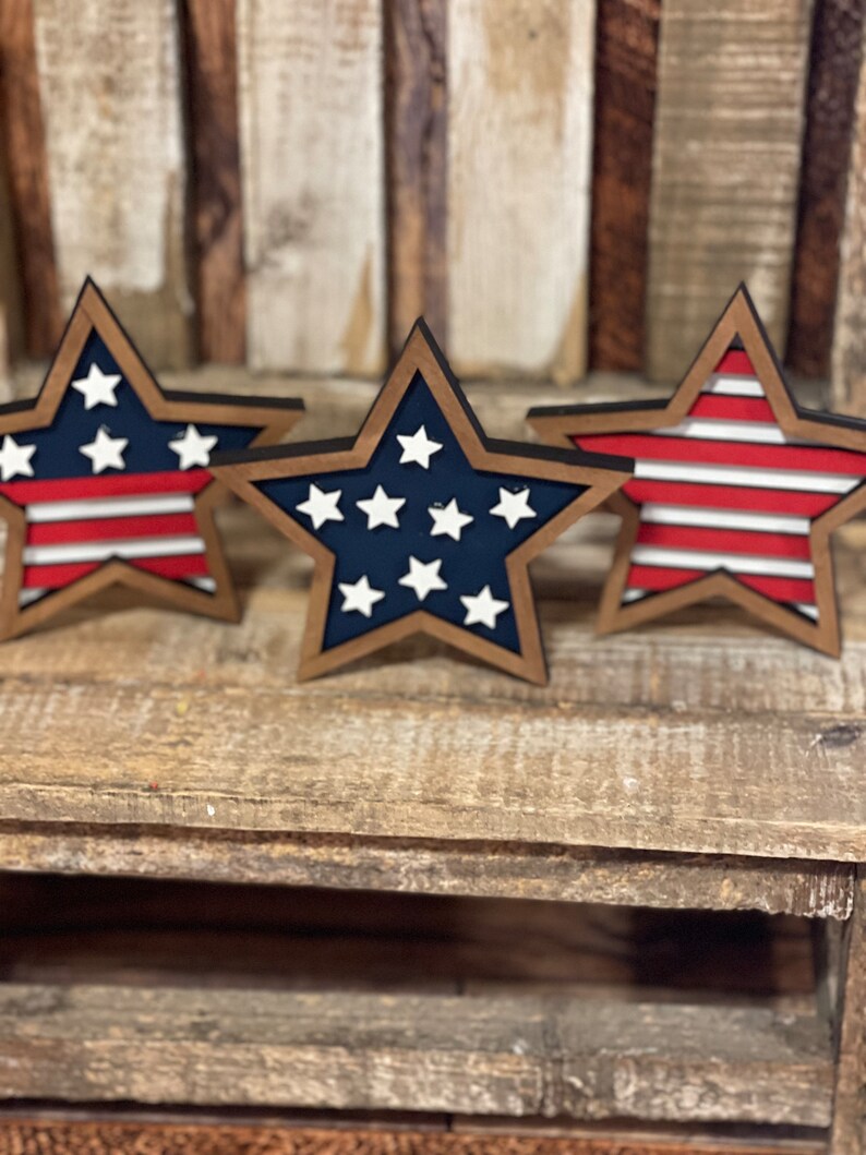 May include: Three wooden stars with a red, white and blue American flag design. The stars are painted in a rustic style and have a brown wood finish.