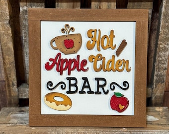 May include: A wooden framed sign with the words "Hot Apple Cider BAR" in colorful lettering. The sign features illustrations of a cup of cider, a cinnamon stick, a donut, and an apple.