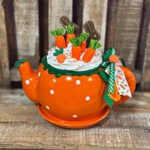 May include: Orange teapot with white polka dots, topped with faux whipped cream, carrot decorations, and brown bunny cutouts. A green and white ribbon with carrot accents adorns the handle. The teapot sits on a matching saucer.