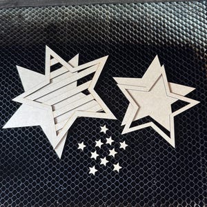 May include: Several unfinished wooden star cutouts of varying sizes are arranged on a dark, textured surface. The stars include layered designs and smaller, individual stars, suitable for crafting or decorative projects.