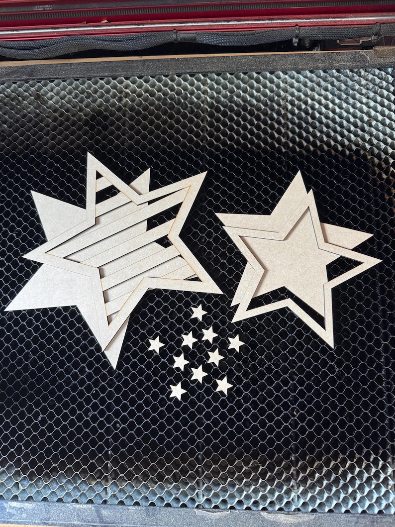 May include: Several unfinished wooden star cutouts of varying sizes are arranged on a dark, textured surface. The stars include layered designs and smaller, individual stars, suitable for crafting or decorative projects.