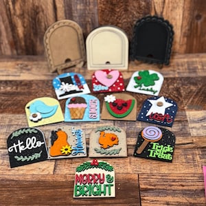 May include: A collection of decorative wooden plaques in various shapes and designs. The plaques feature themes like a bird, ice cream, watermelon, and holiday greetings such as "Hello" and "Merry & Bright". The color palette includes black, white, red, and blue.