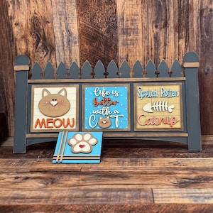 May include: A wooden cat-themed sign with a gray picket fence design. The sign features three panels with cat-related phrases and images, including a cat face, paw print, and the words "Meow," "Life is better with a Cat," and "Spoiled Rotten Catnip."