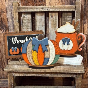 May include: A set of three wooden decorative items including a rectangular sign with the word thankful, a pumpkin with a blue bow and patchwork patterns, and an orange mug with whipped cream and cinnamon sticks, all featuring various fall colors.
