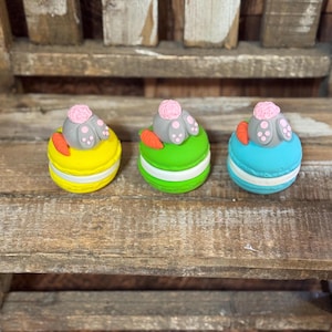 May include: Three colorful Easter-themed macarons. Each macaron has a different color: yellow, green, and blue. They are decorated with a bunny's rear end and a small carrot. The macarons are sitting on a wooden surface.