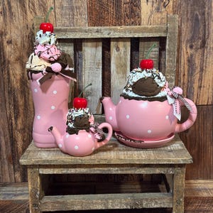 May include: A tall pink polka dot milkshake glass, a small pink polka dot teapot, and a large pink polka dot teapot, all topped with faux whipped cream, chocolate drizzle, and bright red cherries, accented with small brown and white ribbons.