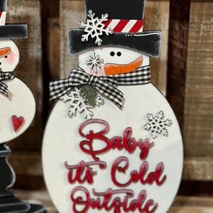 May include: A white wooden snowman figurine with a black top hat, black and white checkered scarf, and red lettering that reads "Baby It's Cold Outside".