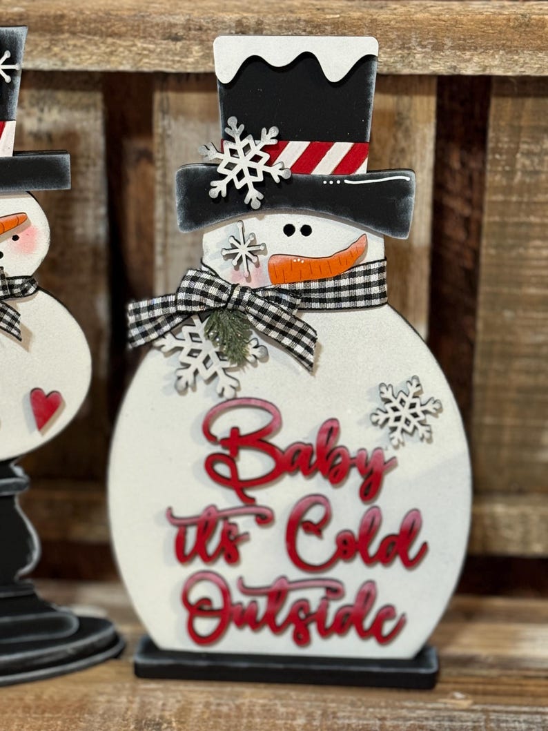 May include: A white wooden snowman figurine with a black top hat, black and white checkered scarf, and red lettering that reads "Baby It's Cold Outside".
