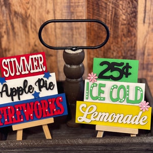 May include: Wooden signs with summer-themed text. One sign reads "SUMMER Apple Pie FIREWORKS" in red, white, and blue. The other sign says "25¢ ICE COLD Lemonade" in green and yellow, with pink flower accents. Both signs are on small easels.
