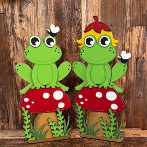 May include: Two wooden frog figurines, each perched atop a red mushroom with white spots. The frogs are green with large eyes and a fly perched on one. The mushrooms sit on a brown base with green grass.