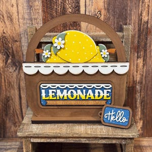 May include: Wooden lemonade stand decoration featuring a brown basket with a handle, a scalloped white trim, and a sign that reads "Lemonade." The basket holds yellow lemons with white polka dots and daisy accents. A small blue sign says "Hello."
