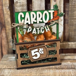 May include: A wooden Carrot Patch sign with a green background and white lettering. The sign sits atop a wooden crate filled with orange carrots. A price tag reads "5¢".