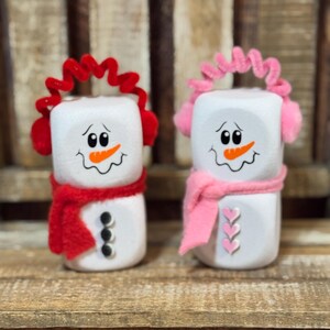 May include: Two snowman-shaped decorations. One snowman wears red earmuffs and a red scarf, the other wears pink earmuffs and a pink scarf. Both have black button eyes, a carrot nose, and a smiling mouth.