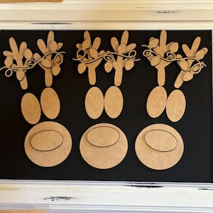 May include: Three unfinished wooden reindeer head cutouts with antlers and holly. The reindeer have two large oval eyes and a smaller oval nose.