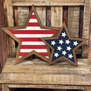 May include: Two wooden star-shaped decorations. One star features red and white stripes, the other has a blue background with white stars. Both have a brown border and are set against a rustic wooden backdrop.